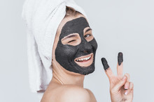 Horizontal Studio Portrait Of Happy Young Woman Blink With Eye With Cosmetic Black Clay Organic Mask On Her Face, Wears Black White On Hair. Female Taking Care Of Face Skin, Isolated On White Wall
