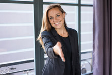 Closeup Portrait Of Smiling Young Beautiful Brown-haired Woman Looking At Camera, Extending Arm For Handshake And Standing At Window. Partnership Concept. Front View.
