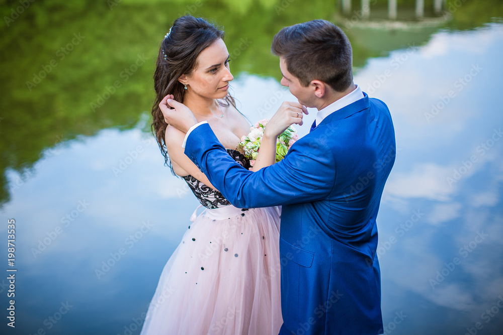 Groom in blue suit straightens bride's hair in pink wedding dress on river bank in park Stock Photo | Adobe Stock Groom in blue suit straightens bride's hair in pink wedding dress on river bank in park Stock Photo | Adobe Stock