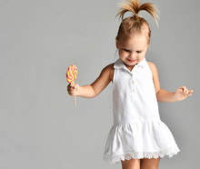 Young Pretty Toddler Girl Kid With Big  Sweet Lollypop Candy In White Dress On Grey