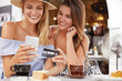© WHstudio Leushin N - Portrait of female models make shopping online, use smart phone, credit card, sit together at cafe interior with aromatic coffee, have positive looks. Best friends recreat together, uses technologies