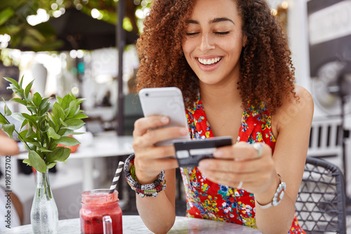Fotografía Beautiful dark skinned young female with cheerful expression, holds smart phone and credit card, banks online or makes shopping while sits against cafe interior
