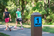 A one mile marker sign post beside a paved pathway with a bright green and sunny background and two woman walking.