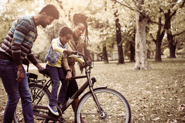  African American mother and father teaching their daughter to drive bike in park.