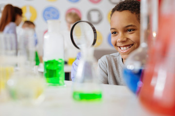 Wall Mural - Great mood in class. Cheerful teenage male student smiling widely while using a magnifying glass and checking the content of a chemical flask