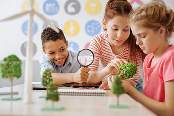 Studying thoroughly. Upbeat teenage students sitting at the table and examining a little tree model while the boy using a magnifying glass for it