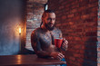 © Fxquadro - A handsome bearded tattoed male with a stylish haircut and muscular body, drinks coffee, leaning on a table.