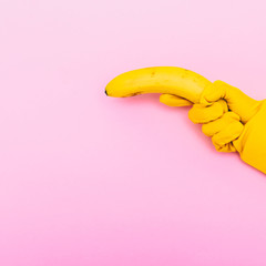 Wall Mural - hand in yellow glove is holding a banana like a gun on a pink background