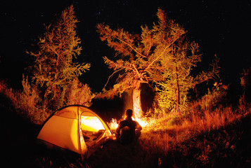 man alone meditative sitting by the camp fire near a travel tent at night in the dark