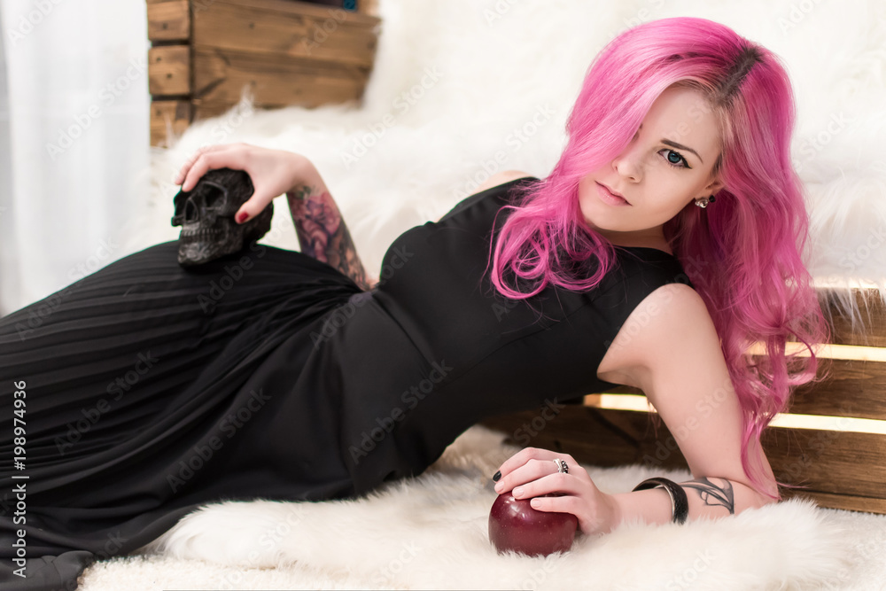 pink hair black dress Beautiful girl with pink hair with a black skull and an apple in her hands.  Informal appearance. Tattoos on the hands. A piercing look. Black dress.  Lying. Stock Photo