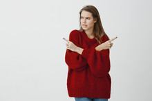 Troubled Girl Making Tough Deicison. Doubtful Attractive Woman In Loose Red Sweater, Crossing Hands And Pointing In Different Directions, Biting Lip Nervously, Wanting Something Over Gray Wall