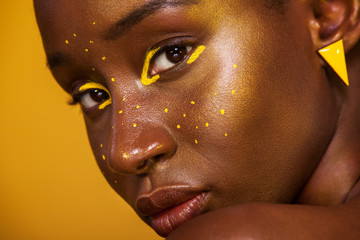 Wall Mural - Closeup portrait of young african woman with yellow makeup on her eyes. Female model laughing against yellow background.