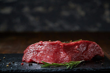 Raw meat beef steaks on black slate board with spices and rosemary over wooden background, copy space