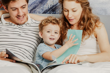 Wall Mural - Happy little boy sitting on bed with young parents, looking through book or magazine, playing with paper and smiling. Woman embracing her husband and reading for son