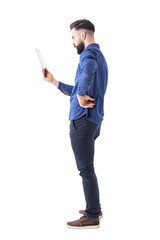 Wall Mural - Profile view of serious bearded businessman holding and looking at tablet computer. Full body isolated on white background. 