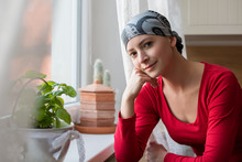 Young Positive Adult Female Cancer Patient Sitting In The Kitchen By A Window, Smiling And Looking At The Camera.