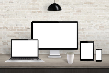 Fototapete - Computer display, laptop, tablet and mobile phone with isolated screen on office desk. White screen for web site design promotion. Brick wall in background.