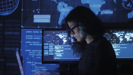 portrait of young woman programmer in eyeglasses working at a computer in the data center filled with display screens