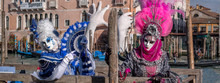 Two women in pink and blue costumes with fans and ornate painted masks at Venice Carnival. Women are standing in front of the Grand Canal with Ca Sagredo in the background