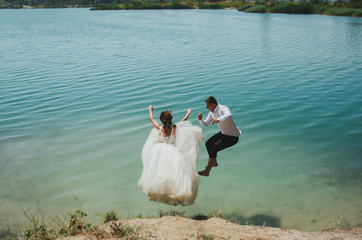 Wall Mural - Wedding couple is jumping into the azure blue lake water. Beautiful bride in puffy dress and groom are having fun. Summer crazy emotions photo on the seaside to ocean coast. Wet wedding clothes.