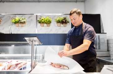 food sale, small business and people concept - male seller wrapping fresh fish into paper at seafood shop