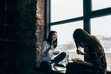 Two cheerful young women dinner sitting windowsill home