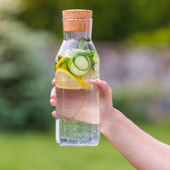 Wall Mural - Female hand holds a bottle with detox drink of cucumber, lemon and mint on a background of green grass