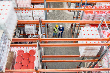 Wall Mural - High angle background image of tall shelves in modern warehouse with two workers wearing hardhats standing in aisle holding clipboard