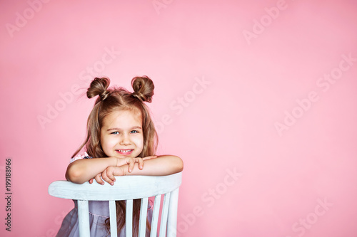 Fotografie, Obraz cute little girl sits on a white chair smiling and looks at the camera on a pink