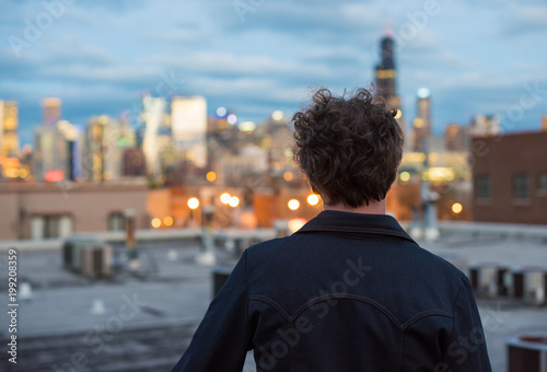 Zdjęcie XXL Atrakcyjny, czterdziestokilkuletni mężczyzna, z długimi, kędzierzawymi brunetkami, na dachu, w mieście, z pejzażem w oddali. Światła zmierzchu i zachodu słońca. Wpatrując się w dal. Kontemplowanie przyszłości.