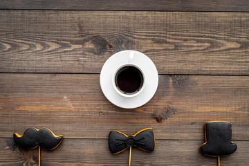 Men's birthday celebration concept. Cookies in shape of moustache, hat, bow tie near coffee on dark wooden background top view copy space