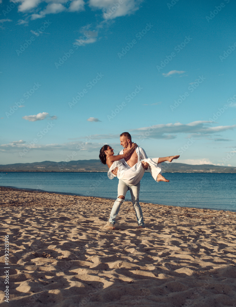 Man carries woman in white dress, couple happy on vacation. Couple in love  stand on beach, seashore. Couple in love having fun, sea and skyline  background. Honeymoon, wedding, just married concept. Stock