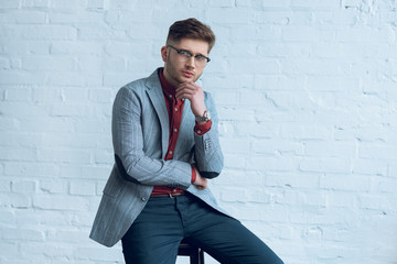 Fototapete - Handsome young man wearing suit and glasses sitting in front of brick wall