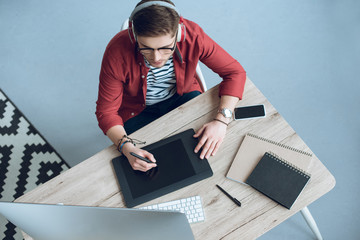 Fototapete - Young man in headphones using graphic tablet by table with computer