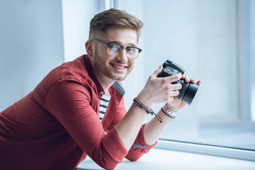Fototapete - Young bearded man in glasses holding camera by window