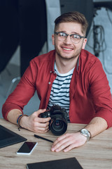 Fototapete - Smiling man wearing glasses and holding camera by table with computer