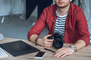 Fototapete - Young bearded man holding camera by table with computer