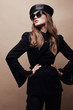 Fashion portrait of beautiful young woman in black flared jacket, black leather beret cap and dark sunglasses with veil. Standing with hands on waist