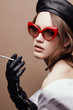© monchak - Fashion portrait of beautiful young woman in leather beret cap, red cat eye retro sunglasses, black leather glove with cigarette in hand