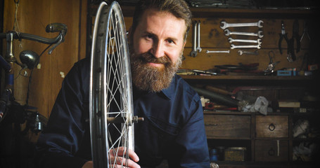 Well-groomed handsome bearded master hipster, specialist in bicycles, repairing a bicycle in his workshop, wheels, frame, spokes, the background of tools. Concept: pro bike, cycle passion, lifestyle.