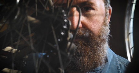 Well-groomed handsome bearded master hipster, specialist in bicycles, repairing a bicycle in his workshop, wheels, frame, spokes, the background of tools. Concept: pro bike, cycle passion, lifestyle.