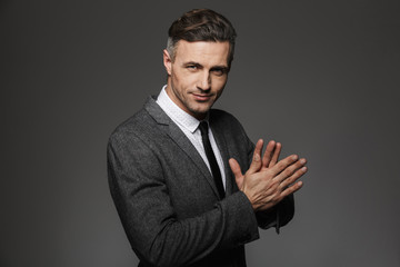 Photo of unshaved successful man 30s wearing office suit and black tie smiling on camera holding hands together, isolated over gray background