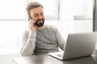 © Drobot Dean - Image of businesslike man 30s wearing casual clothing working on laptop, while sitting at table near window indoor and speaking on cell phone