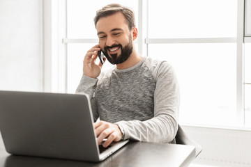 Sticker - Image of pleased guy 30s wearing casual clothing using laptop and talking on cell phone, while sitting at home workplace