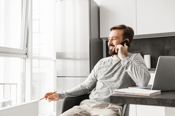 Image of businesslike man 30s wearing casual clothing working on laptop, while looking out window and speaking on cell phone at home