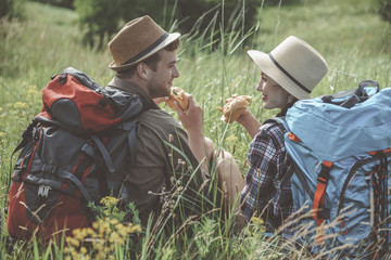 Lunch. Positive young couple with backpacks is sitting on the grass and holding appetizing sandwich while looking at each other with love. Back view