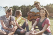 © Yakobchuk Olena - Calm people sitting on grass. Concentrated man performing song on guitar while companions listening and smiling. Focus on woman looking at camera with joy