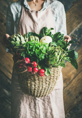 Female farmer in linen apron holding basket of fresh garden vegetables and greens in her hands, rustic wooden barn wall at background. Local market or organic produce concept