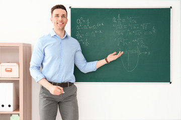 Young male teacher explaining math formulas written on blackboard in classroom
