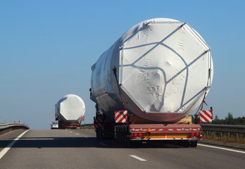 Two trucks trucks with a police escort car carrying oversized cargo on the asphalt road in the summer against the blue sky